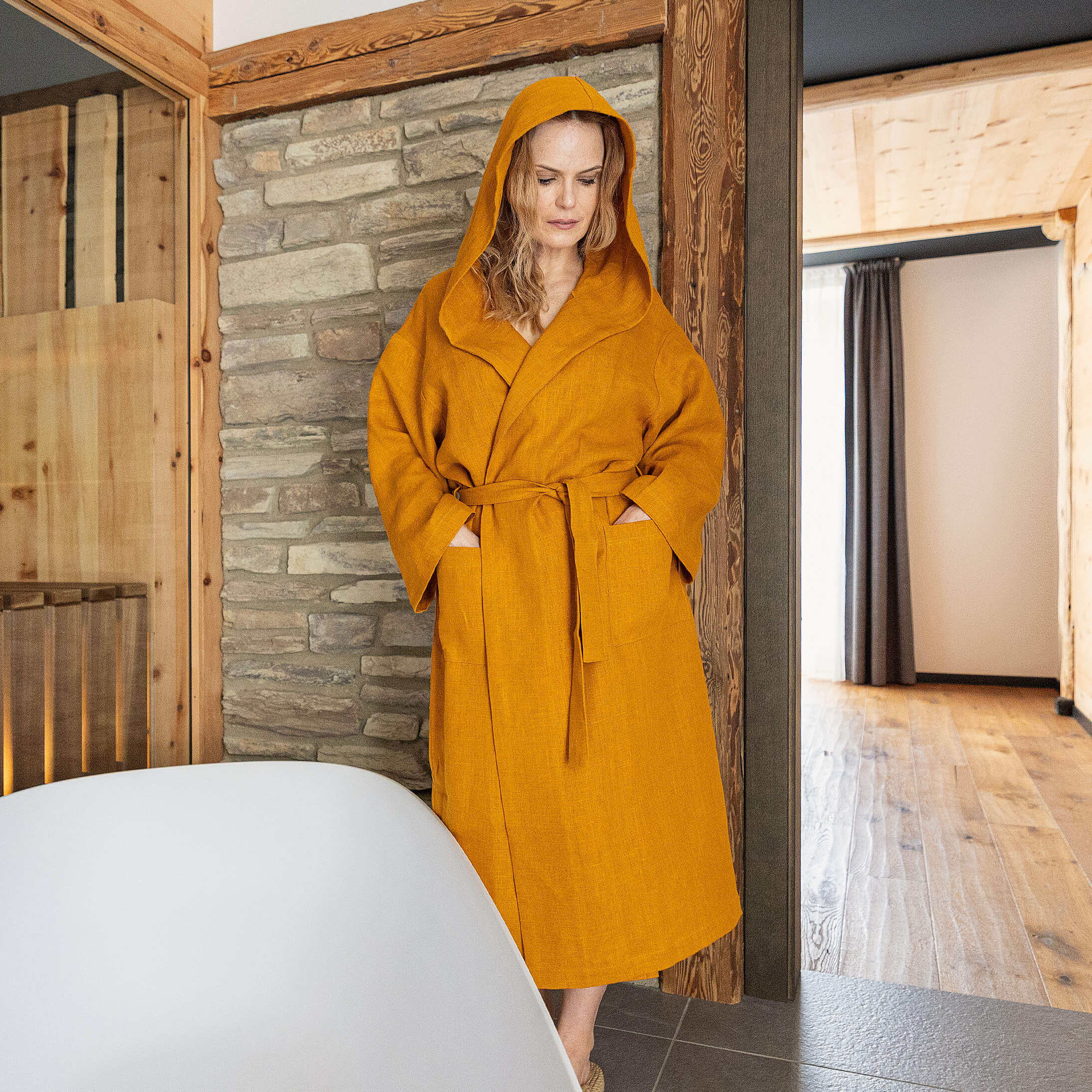 Woman in a amber yellow robe standing in a modern bathroom with stone walls.