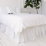 Bright White bed with ruffled bedding and a plant in the corner on a white wooden floor.