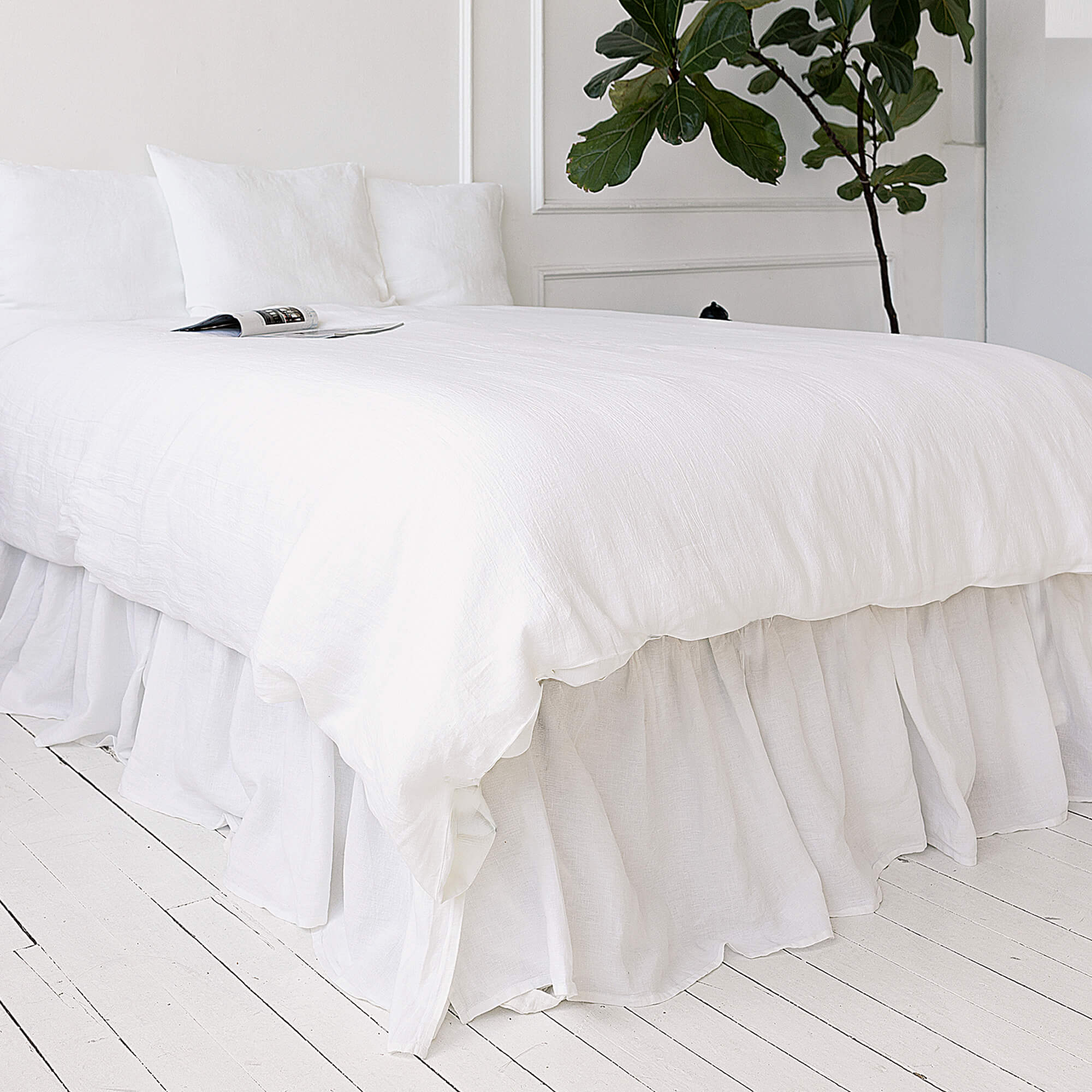Bright White bed with ruffled bedding and a plant in the corner on a white wooden floor.