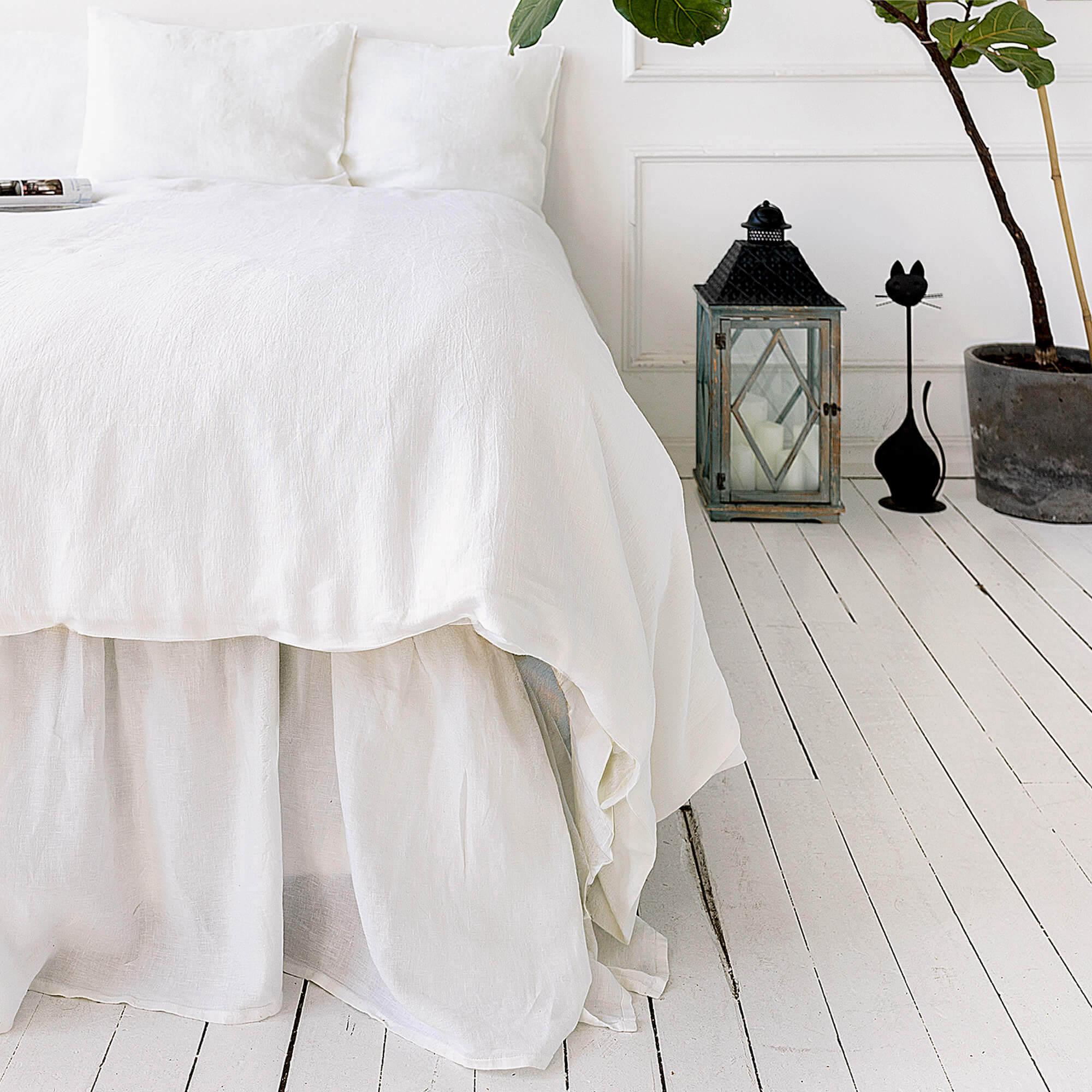 Bright White bed with a white duvet on a white wooden floor, with a lantern and plant in the background.