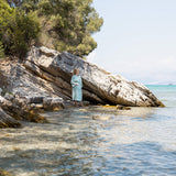 Woman in a duck egg blue bathrobe standing on a rocky beach with trees and clear water.
