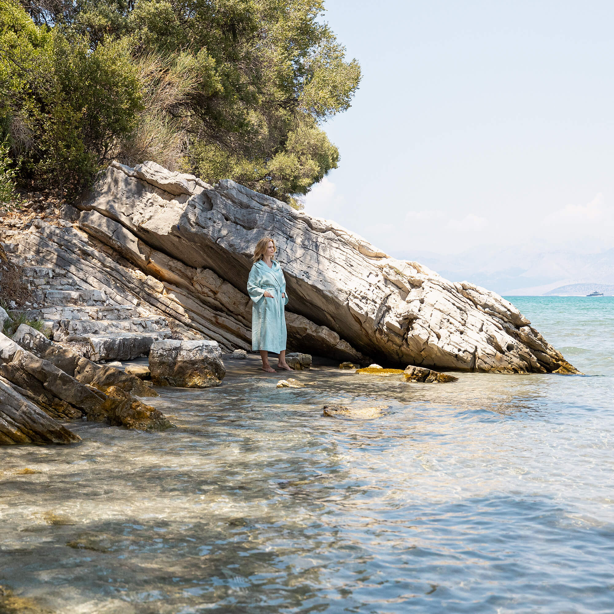 Woman in a duck egg blue bathrobe standing on a rocky beach with trees and clear water.