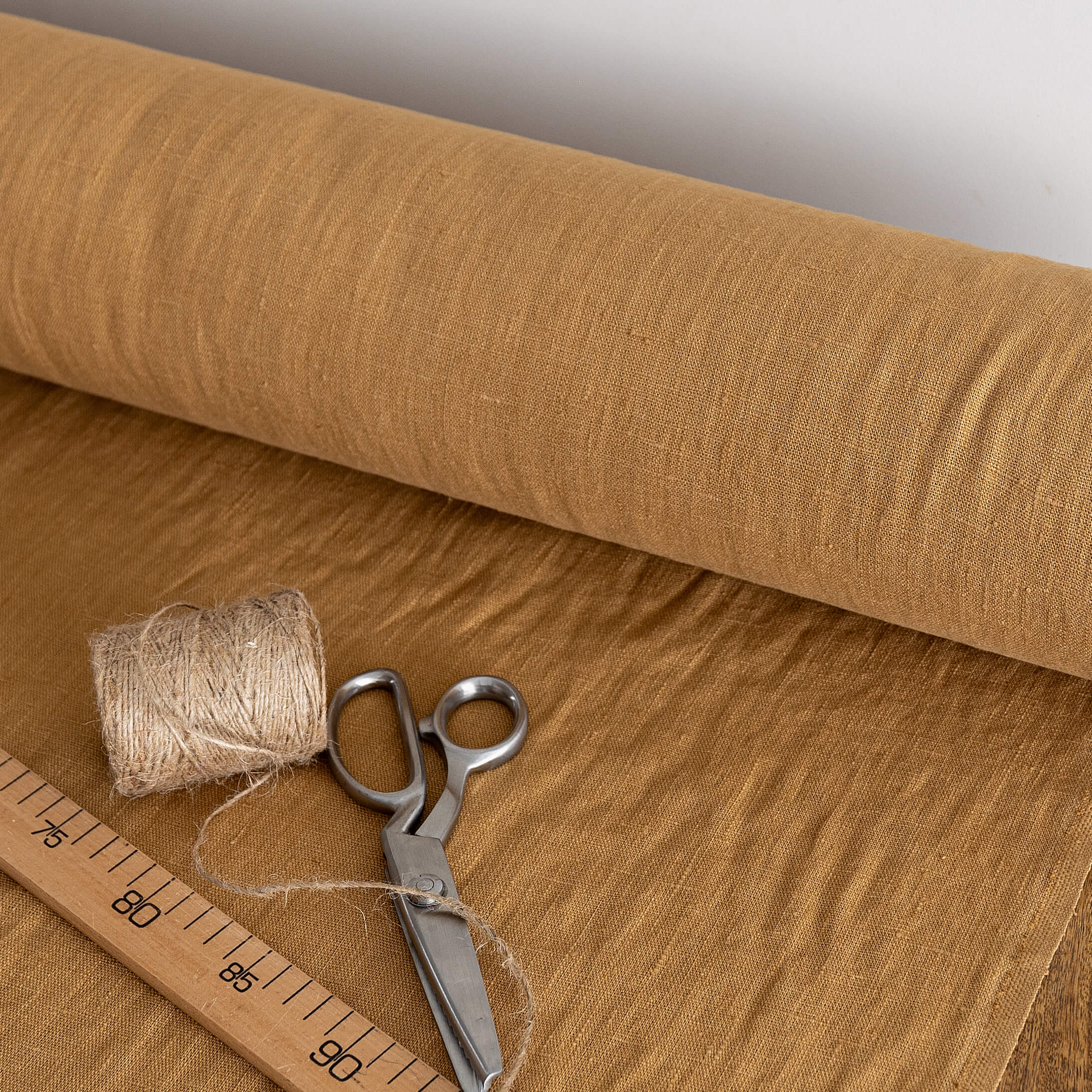 Roll of dusty mustard fabric with scissors, thread, and a ruler on a wooden surface.