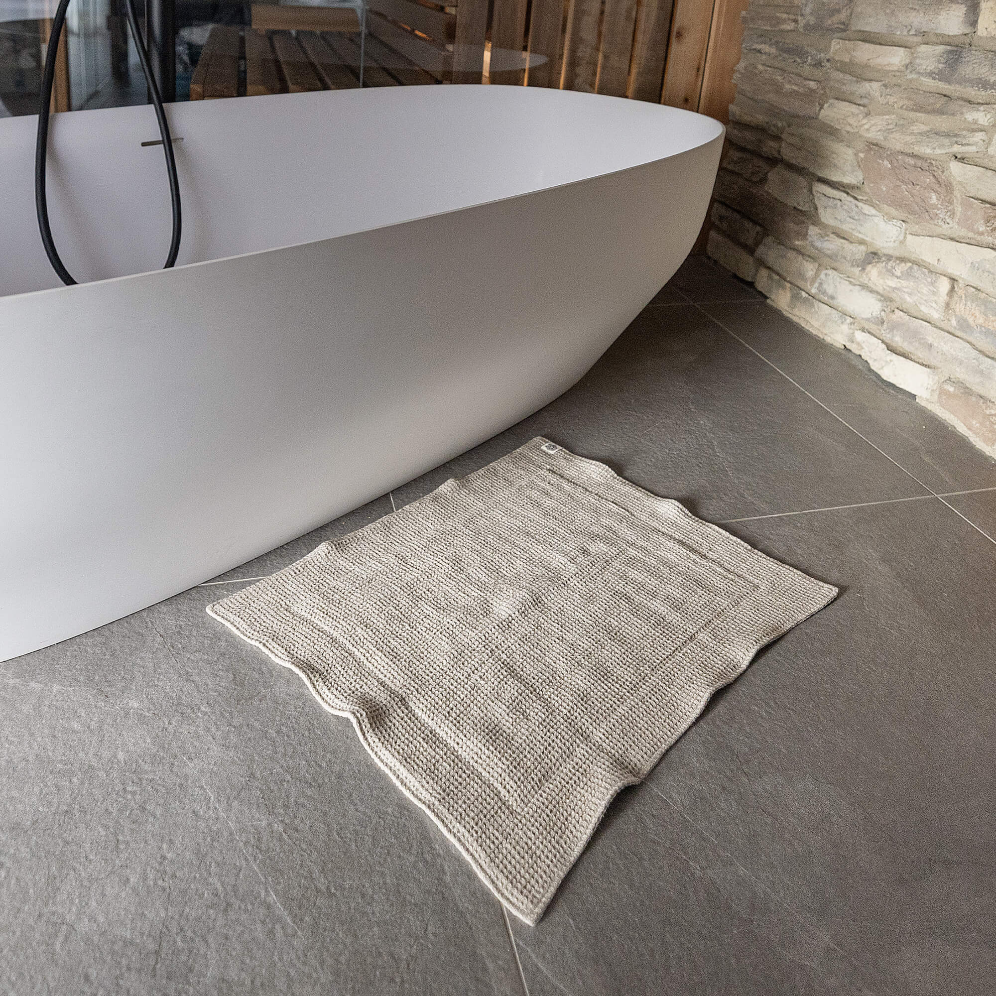 Gray bath mat on a tiled floor next to a white freestanding bathtub in a bathroom.