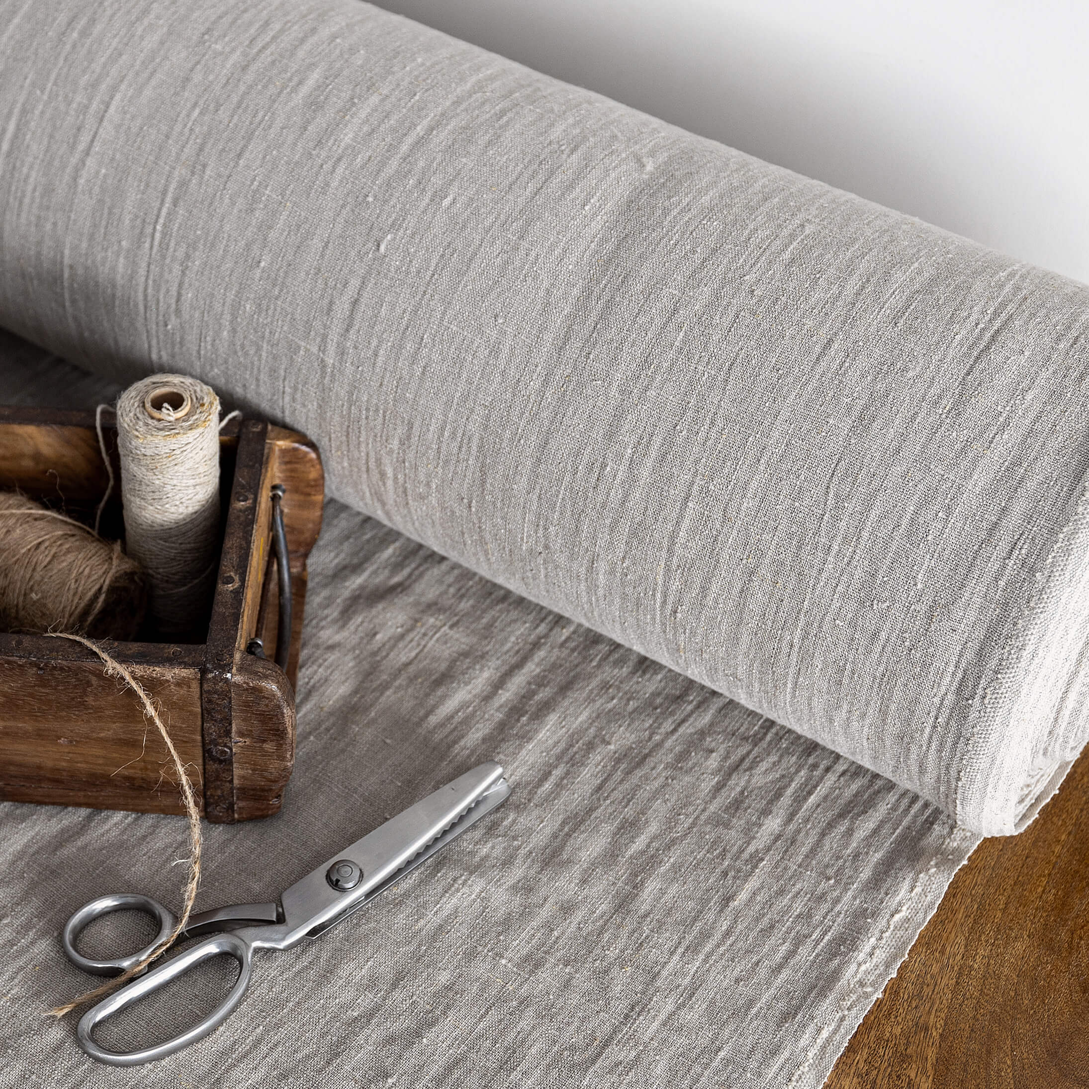 Roll of unbleached linen fabric with a wooden box containing thread and scissors on a wooden surface.