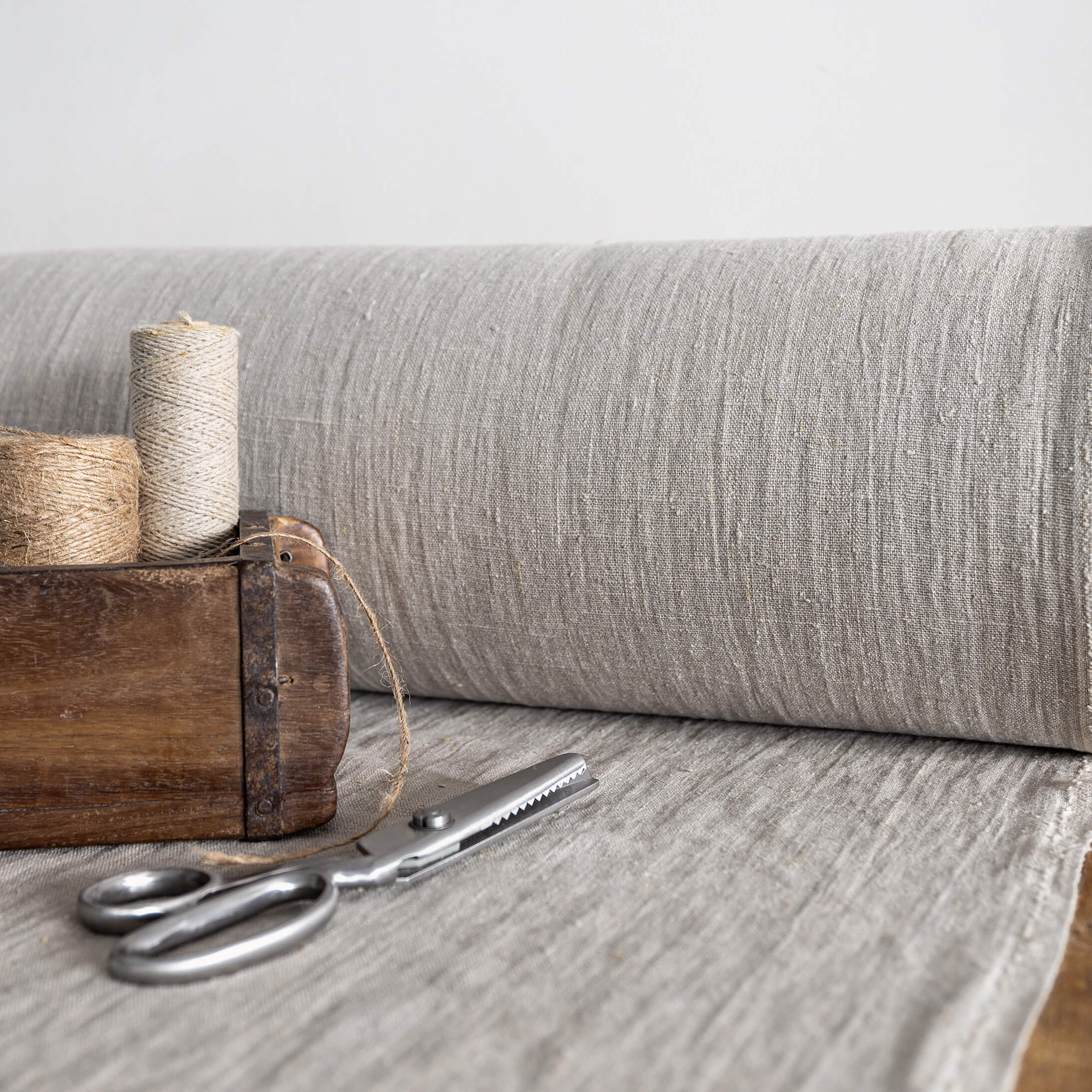 Roll of unbleached linen fabric with a wooden box containing twine and scissors on a light wooden surface.
